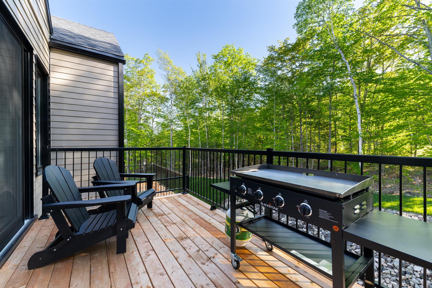 Terrasse en bois avec chaises noires et barbecue, entourée d'arbres verts.