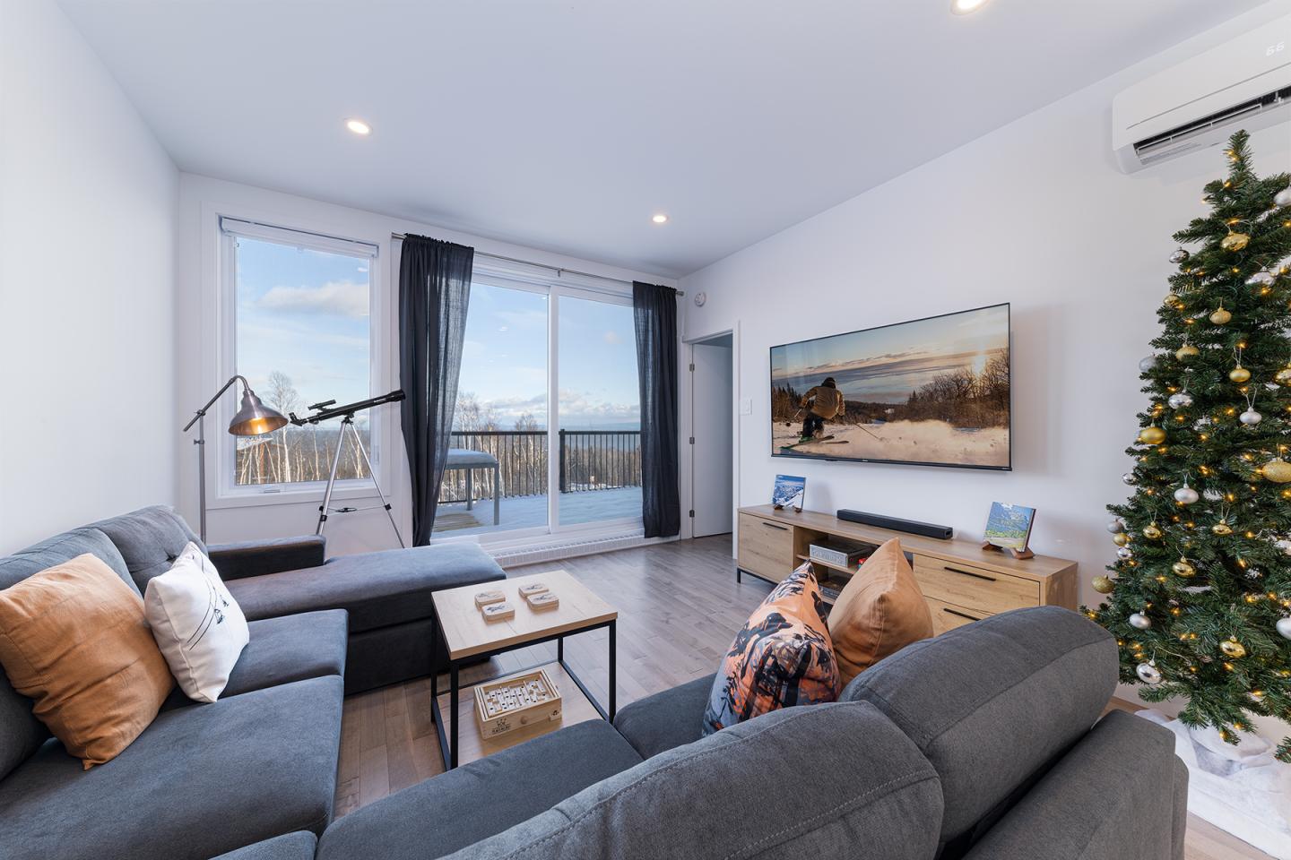 Salon moderne avec canapé gris, sapin de Noël et grande fenêtre sur balcon.