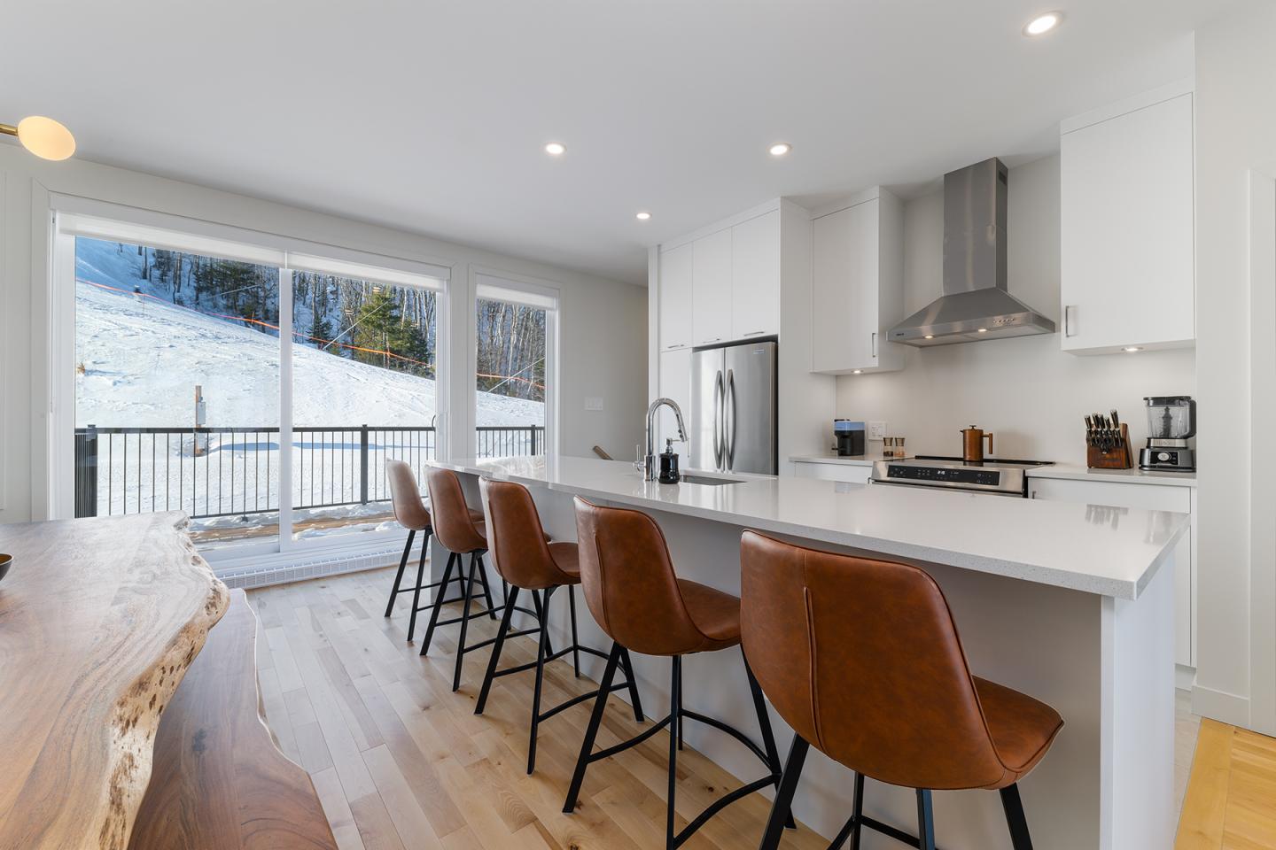 Cuisine moderne avec comptoir blanc, chaises en cuir marron et vue sur un paysage enneigé.