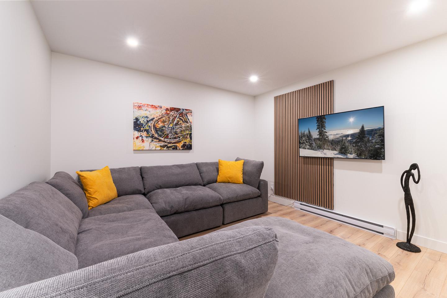 Salon moderne avec canapé gris, coussins jaunes et téléviseur mural.