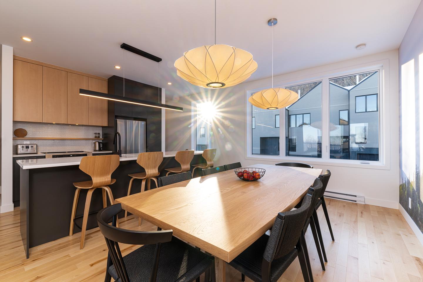 Cuisine moderne avec grande table en bois, chaises noires, et lumière naturelle.