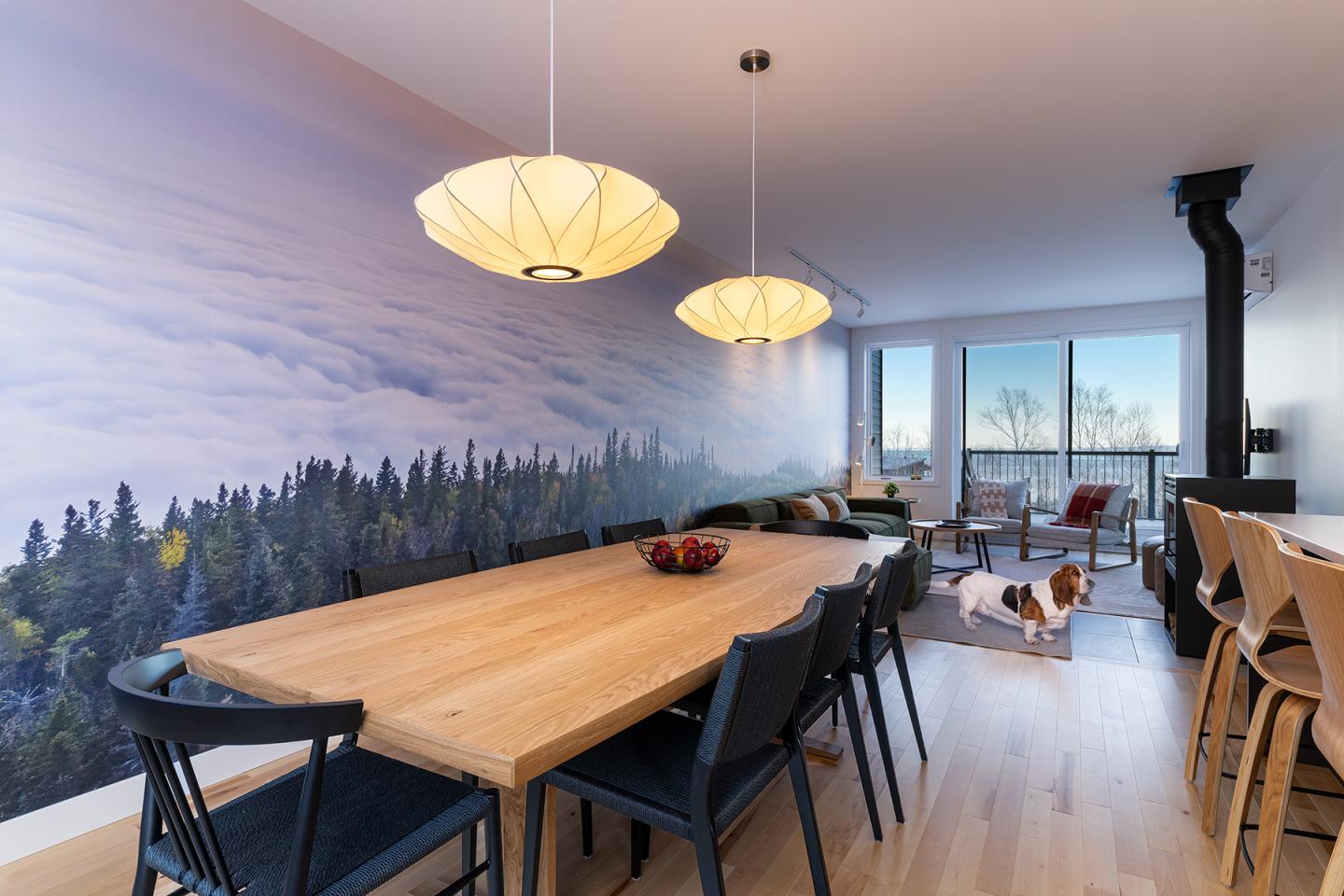 Salle à manger moderne avec une grande table en bois, mur forêt, et un chien près des fenêtres.