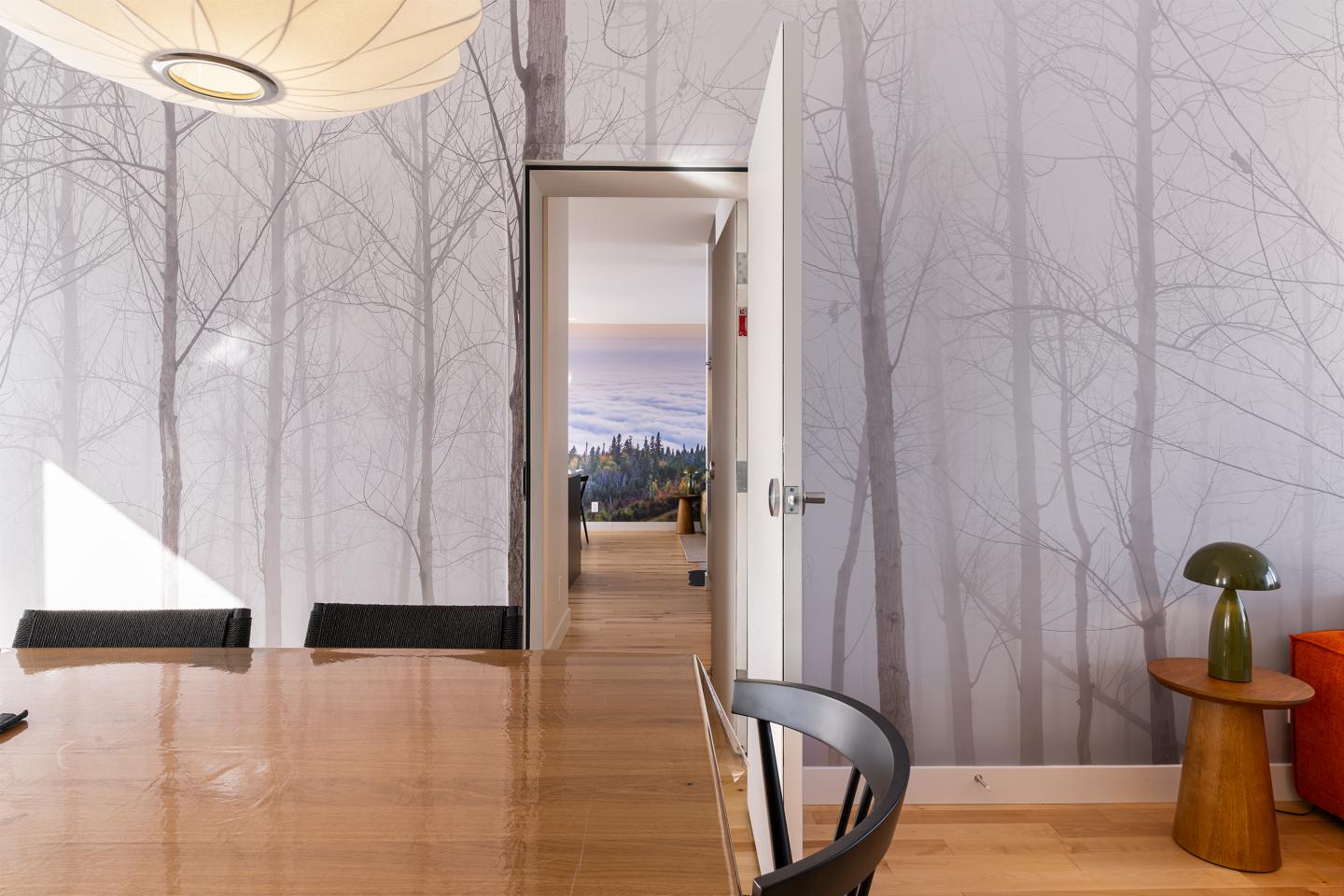 Salle à manger moderne avec mur à motif d'arbres, vue sur un paysage à travers la porte.