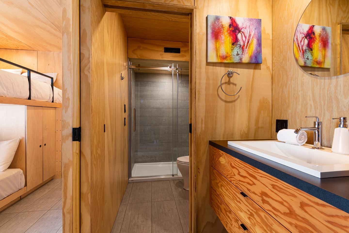 Salle de bain moderne en bois, douche vitrée, lavabo et tableaux colorés.