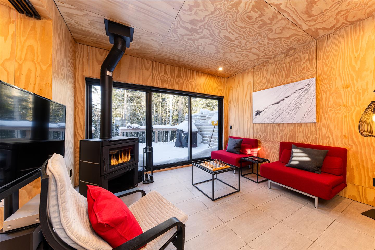 Salon moderne avec poêle à bois, canapé rouge, grande baie vitrée, vue sur la neige.