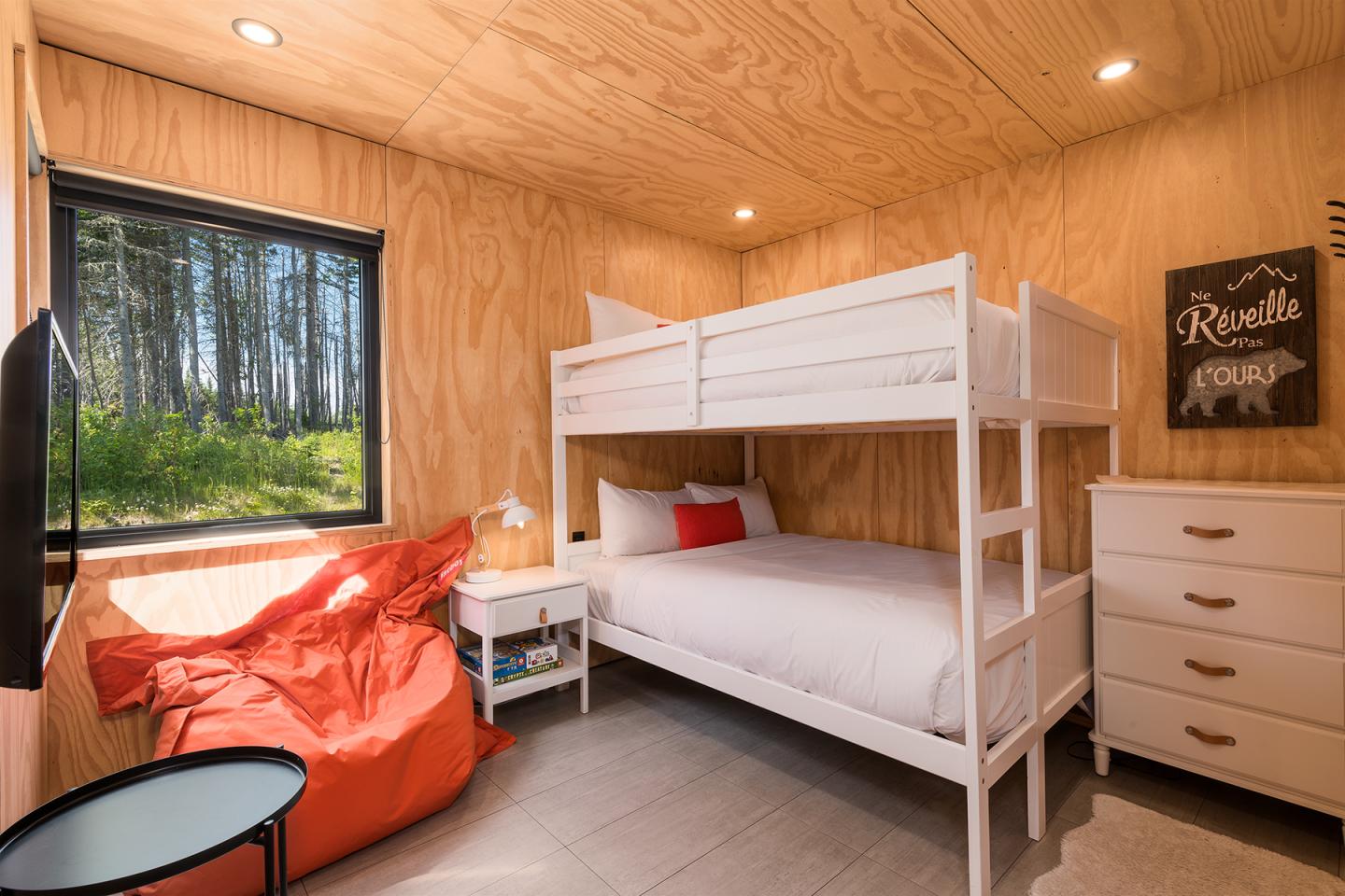 Chambre en bois avec lits superposés, chaise orange et vue sur la forêt par la fenêtre.