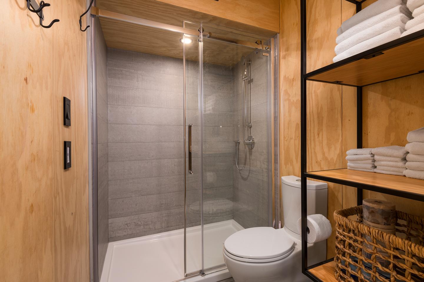 Salle de bain moderne avec douche vitrée, toilettes et étagères en bois.