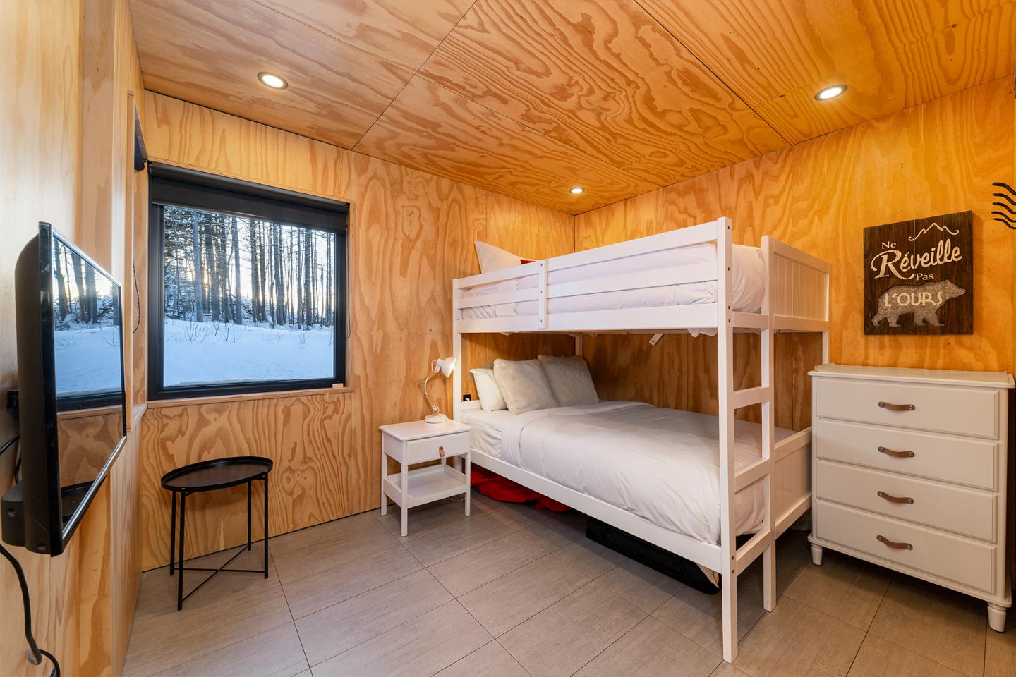 Chambre en bois avec lits superposés blancs et vue sur la neige par la fenêtre.