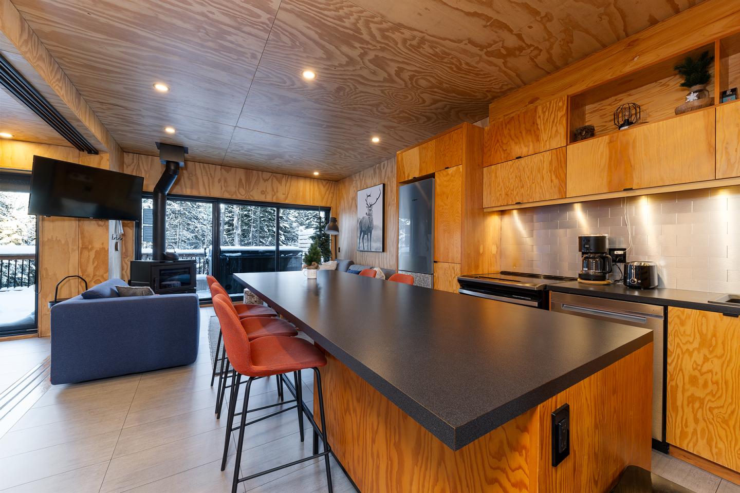 Cuisine moderne en bois avec comptoir noir, tabourets rouges, et grandes fenêtres.
