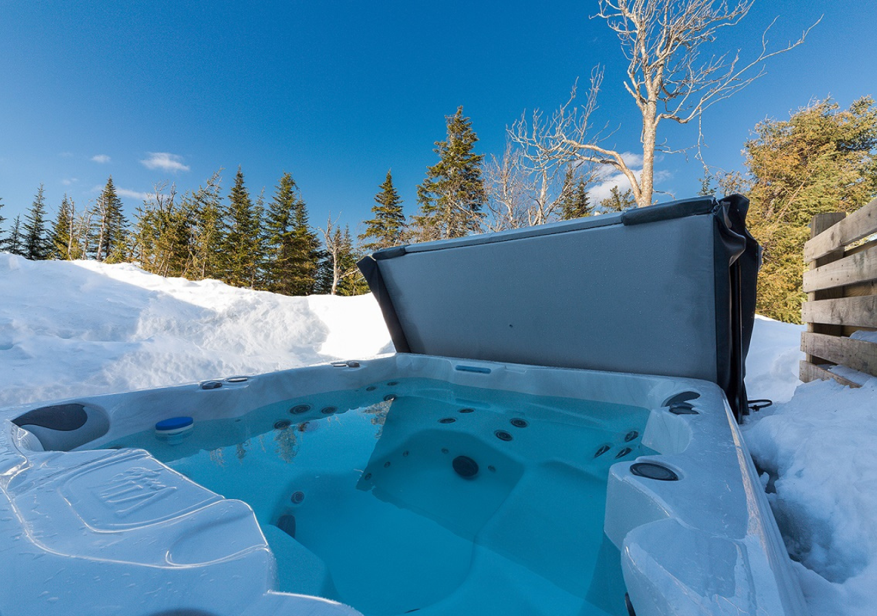 Jacuzzi extérieur enneigé sous un ciel bleu, entouré de sapins.