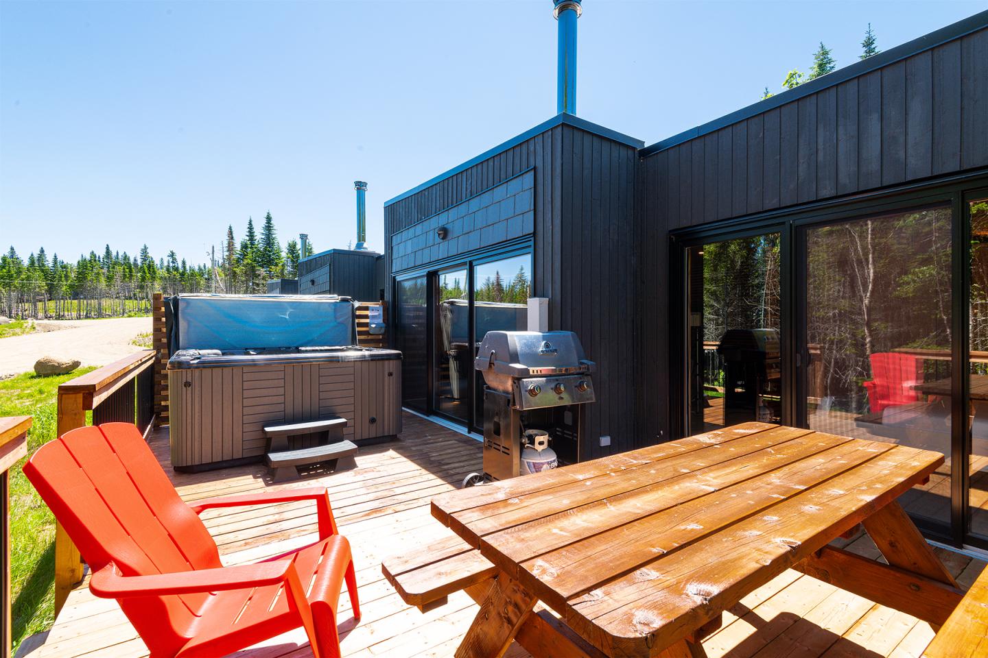 Terrasse en bois avec bain à remous, barbecue, chaises rouges et table, sous un ciel bleu.