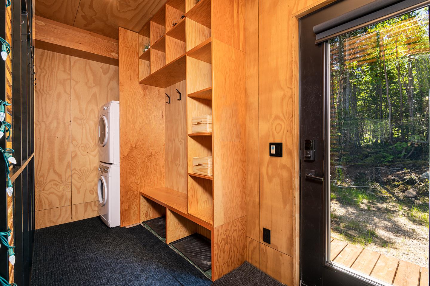 Intérieur en bois d'une entrée avec étagères, lave-linge et vue sur la forêt.