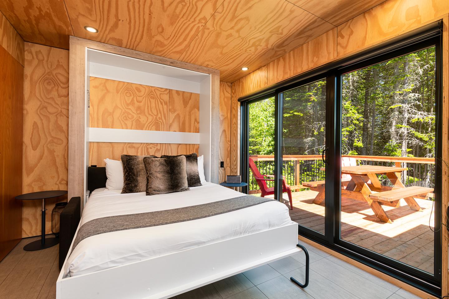 Chambre en bois avec lit, baies vitrées donnant sur terrasse ensoleillée et forêt.