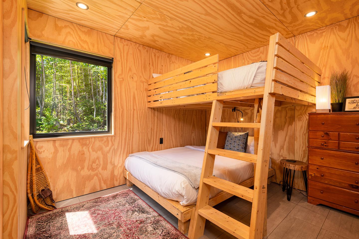 Chambre en bois clair avec lit superposé, fenêtre sur verdure.
