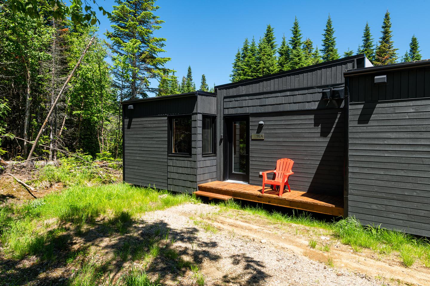 Petite maison noire en bois, chaise orange sur la terrasse, entourée de forêt.