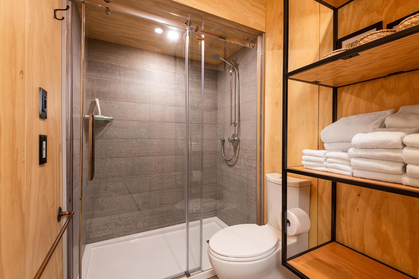 Salle de bain moderne avec douche vitrée, toilettes et étagères en bois.