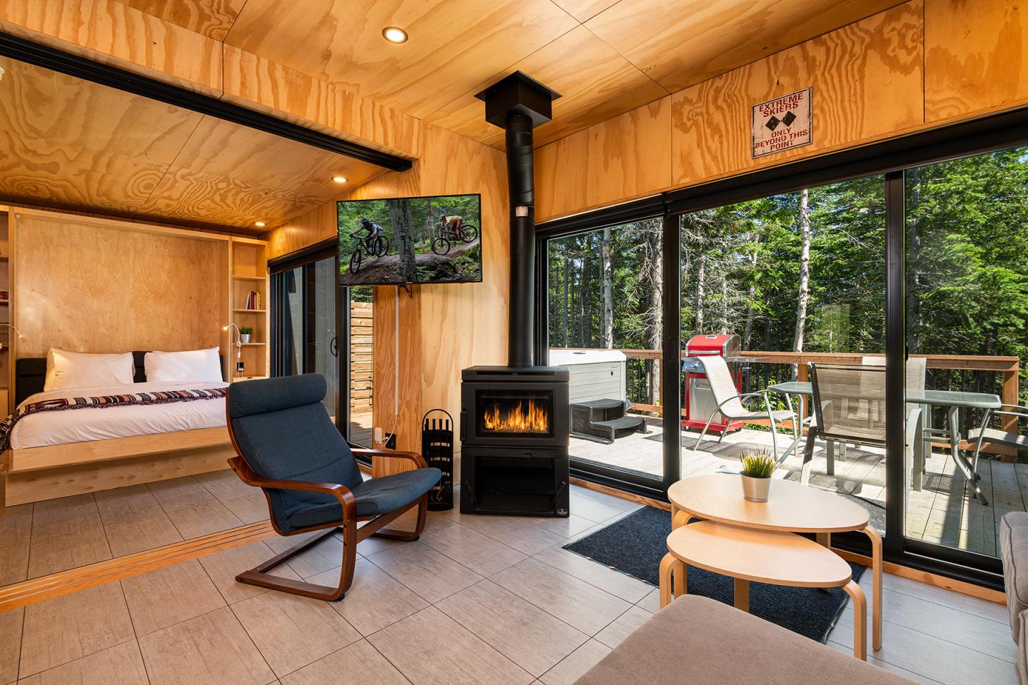 Salon cosy en bois avec fauteuil, table basse, poêle et vue sur terrasse et forêt.