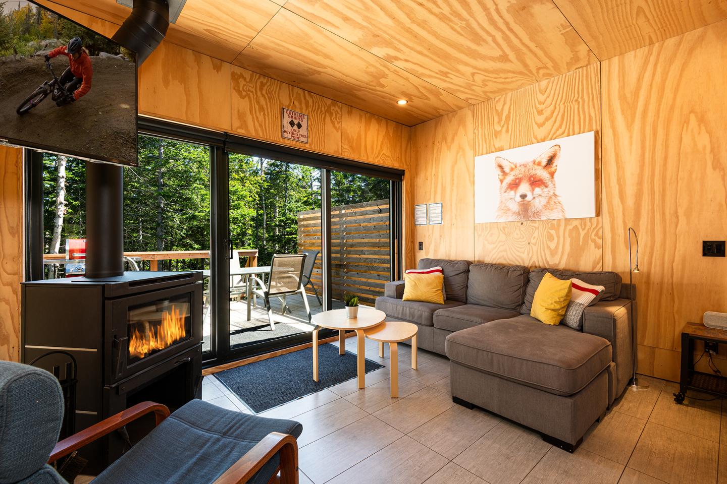 Salon moderne avec poêle, canapé gris, et affiche de renard. Vue sur terrasse boisée.