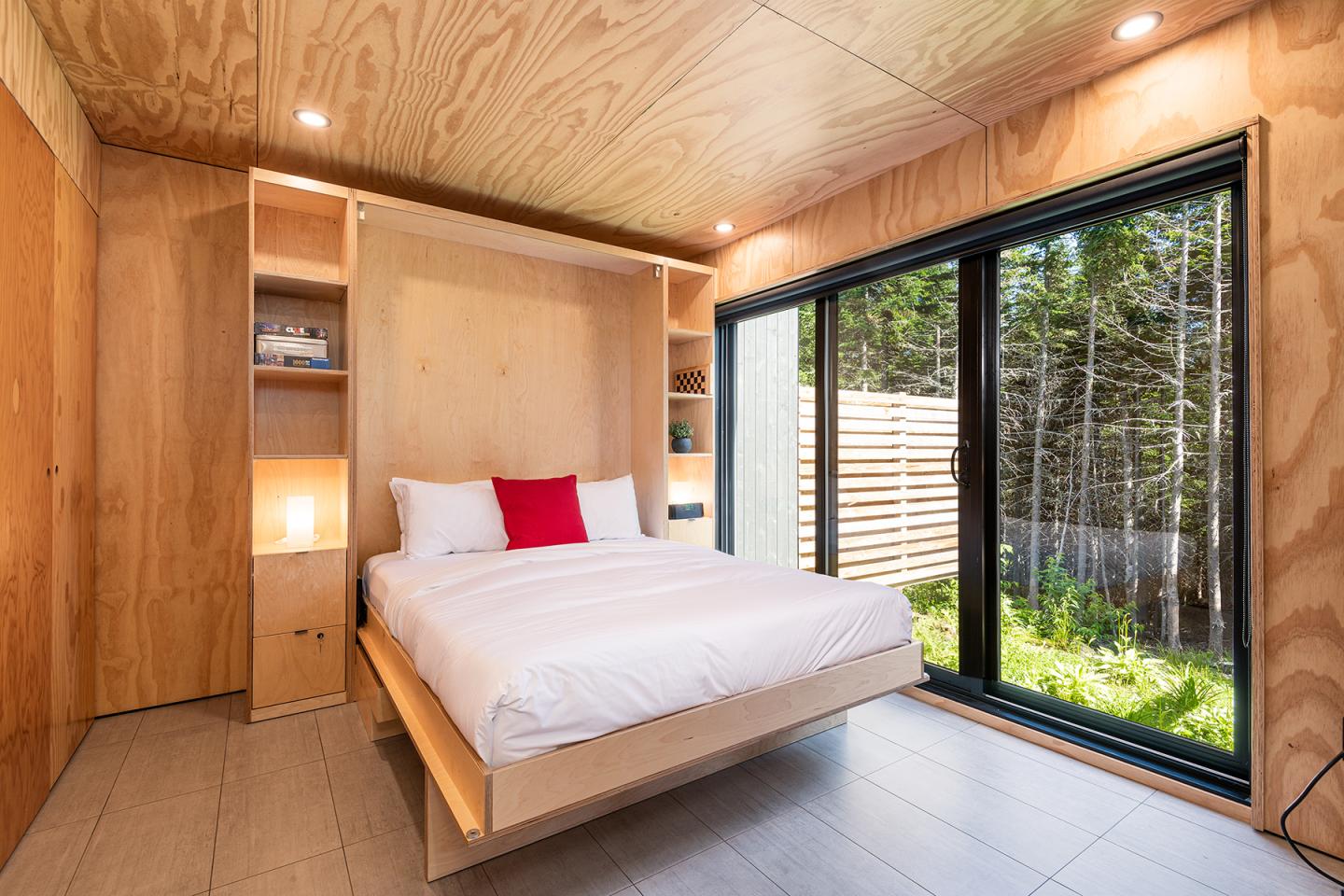 Chambre moderne en bois avec fenêtre, lit à coussin rouge, vue forêt.