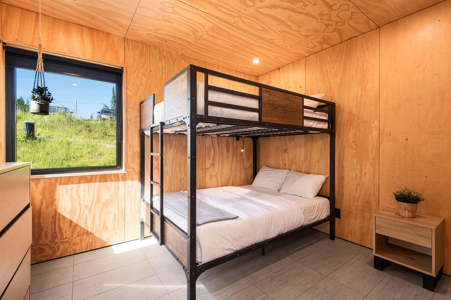 Chambre en bois avec lits superposés, fenêtre donnant sur la verdure.