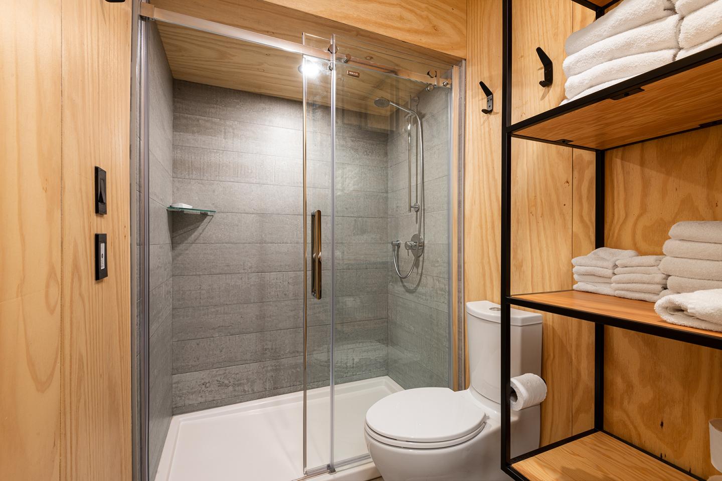 Salle de bain moderne avec douche vitrée, étagères en bois et toilettes blanches.