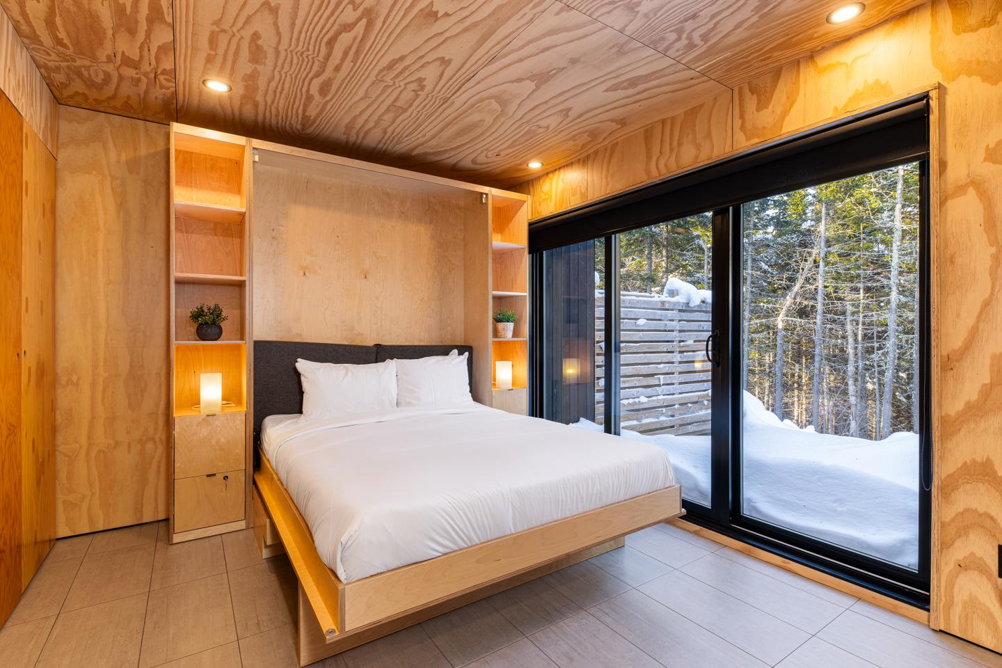 Chambre moderne en bois clair avec lit double, grande baie vitrée donnant sur forêt enneigée.