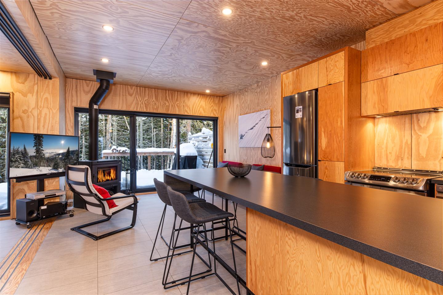 Cuisine moderne en bois, comptoir long, poêle à bois, et vue sur la forêt.