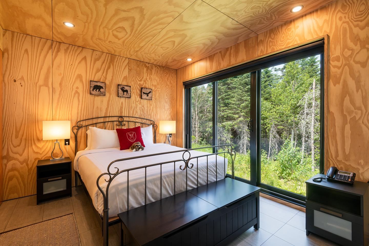 Chambre en bois avec lit, grandes fenêtres donnant sur la forêt.