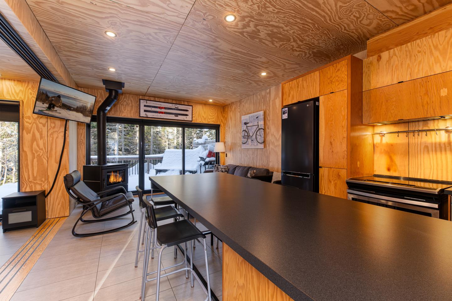 Grand salon en bois avec poêle à bois, grandes fenêtres et table longue noire.