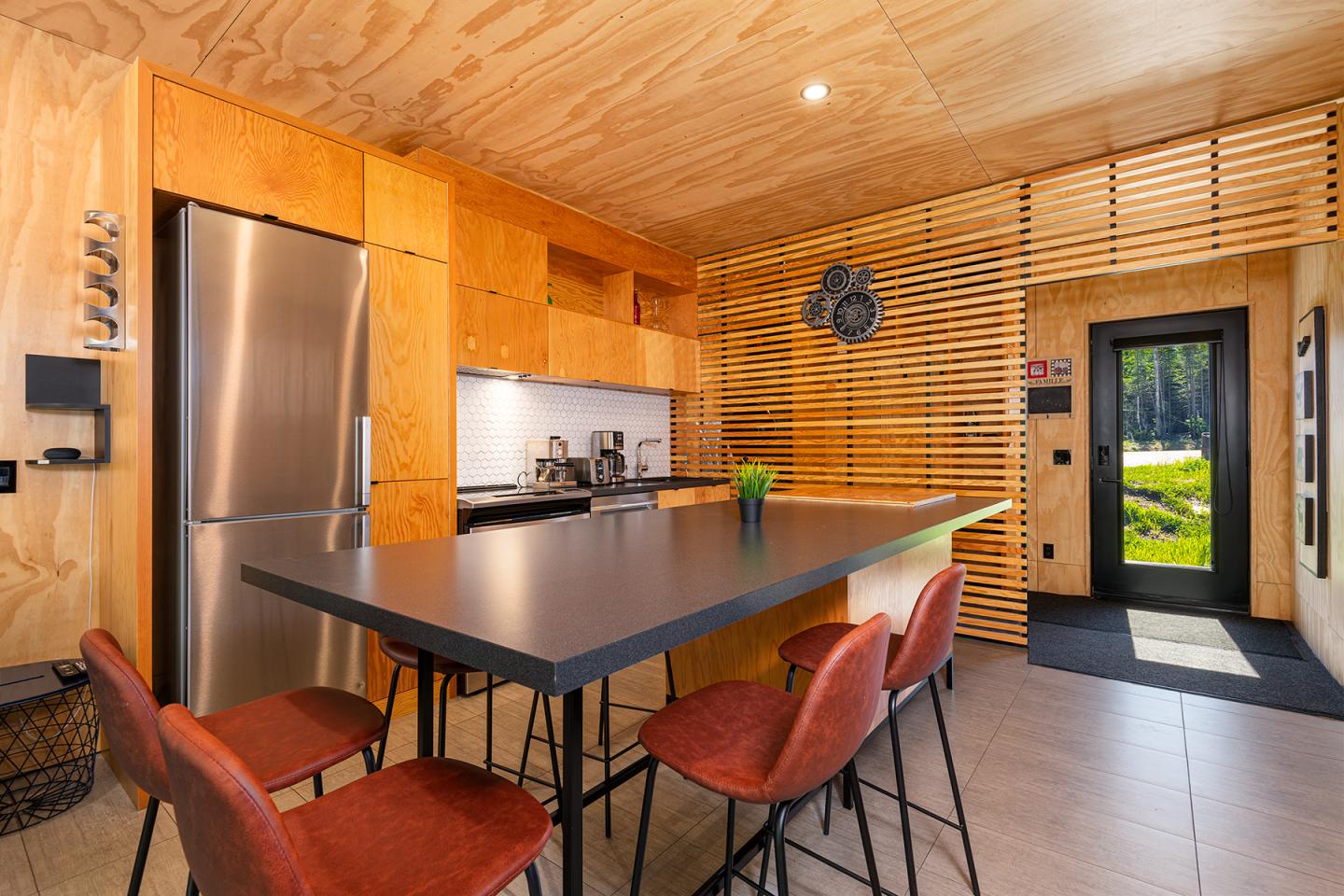 Cuisine moderne en bois, comptoir noir, chaises rouges, réfrigérateur inox, grande fenêtre.