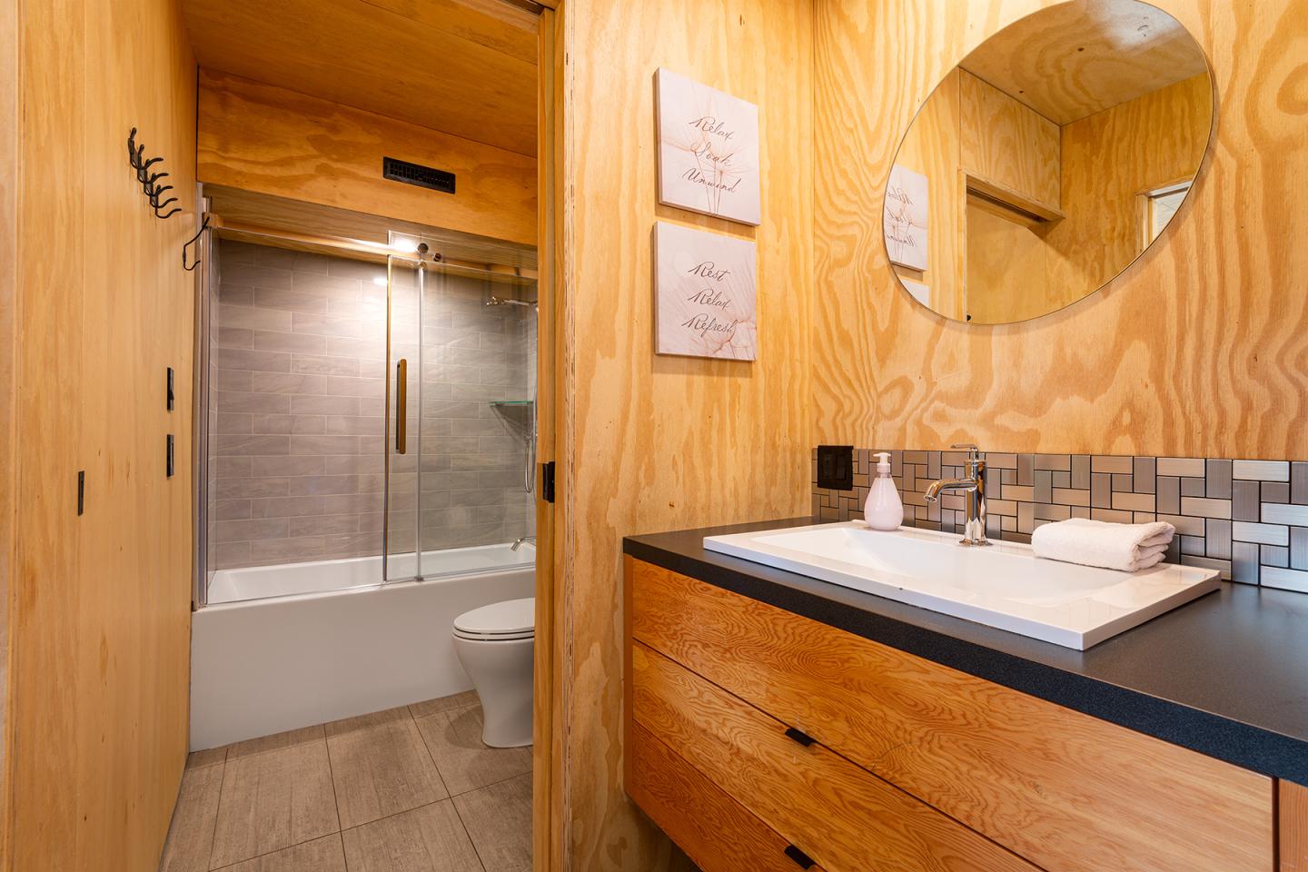Salle de bain en bois clair, miroir rond, lavabo et baignoire moderne.