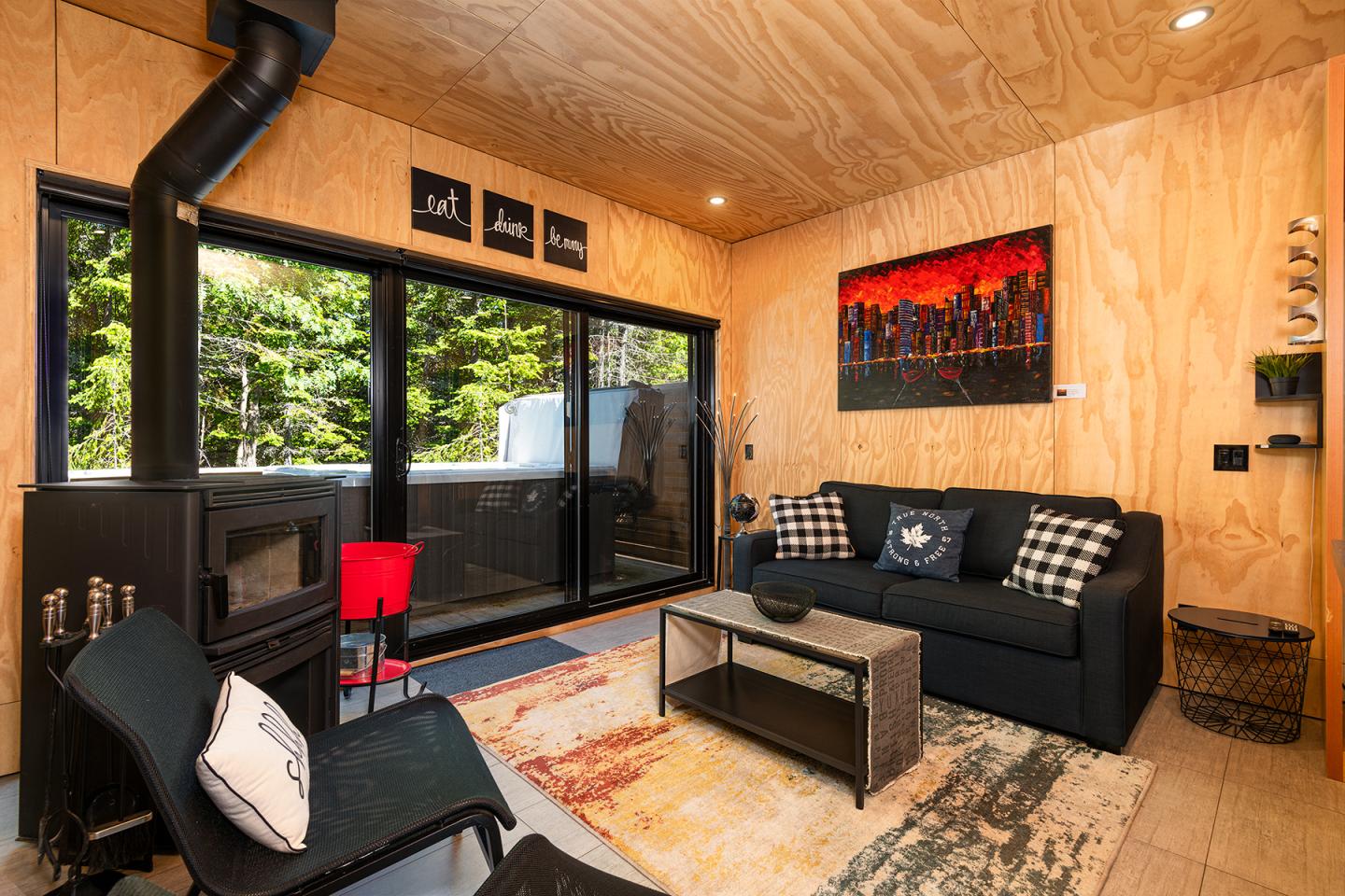 Salon moderne avec murs en bois, canapé noir, poêle à bois, grandes fenêtres sur jardin.
