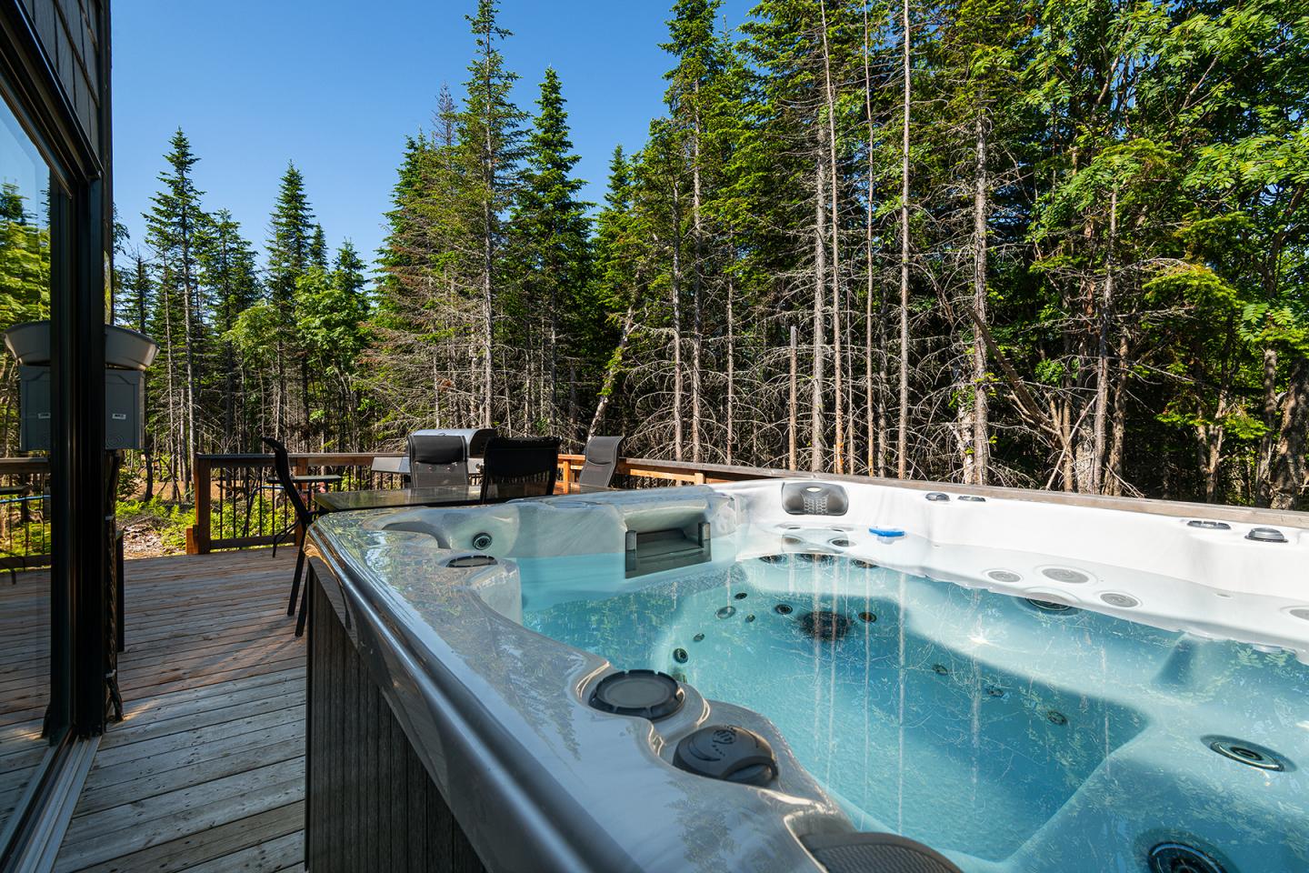 Jacuzzi sur une terrasse en bois, entouré de forêts sous un ciel bleu.