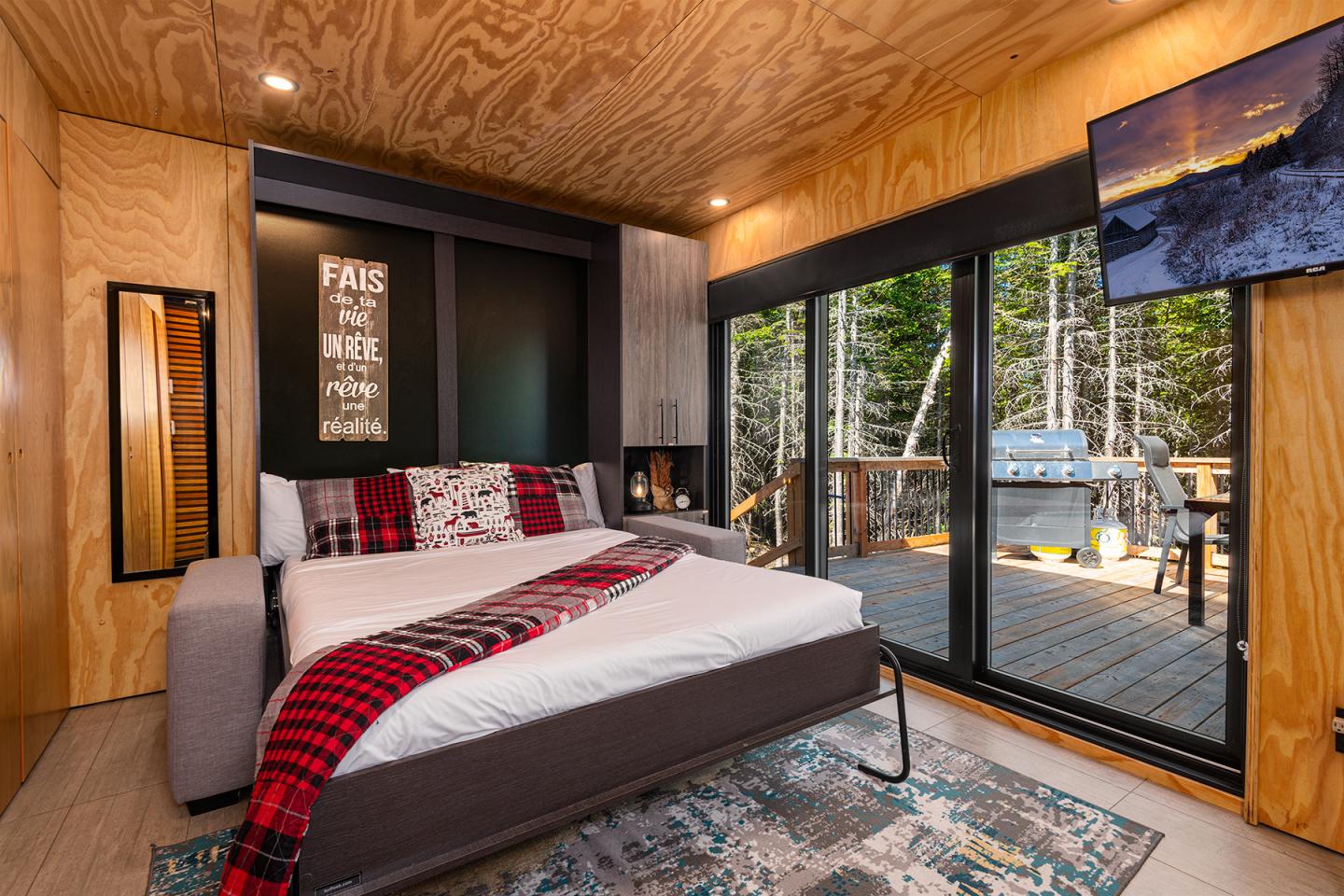 Chambre moderne avec lit, plaid rouge, grande baie vitrée donnant sur terrasse boisée.