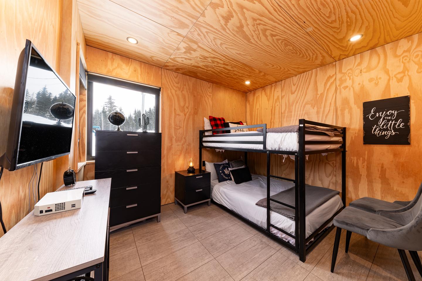 Petite chambre avec lit superposé, bois clair, télé, commode et fenêtre.