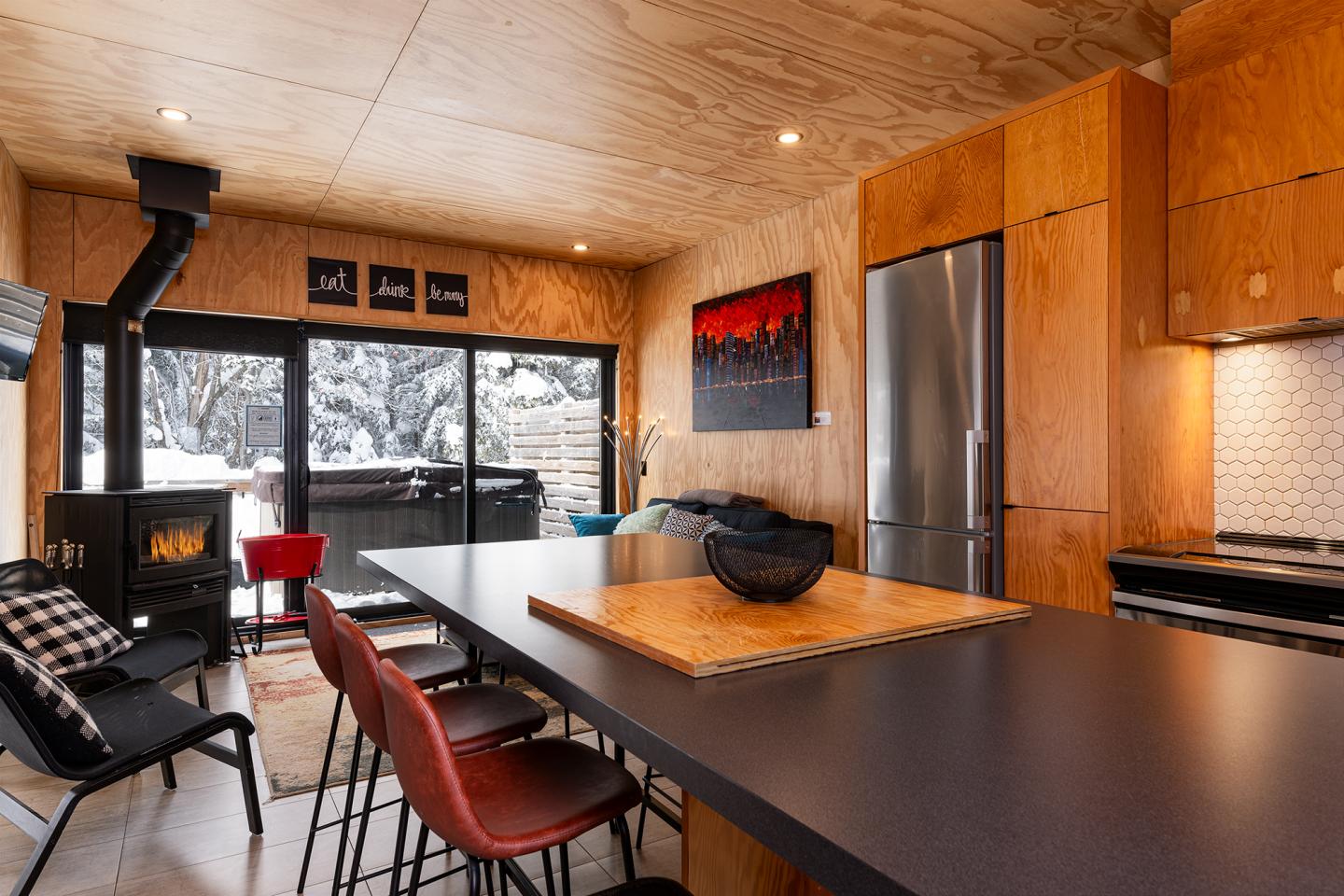 Cuisine moderne en bois avec cheminée, grande table noire, et fenêtre donnant sur un paysage enneigé.