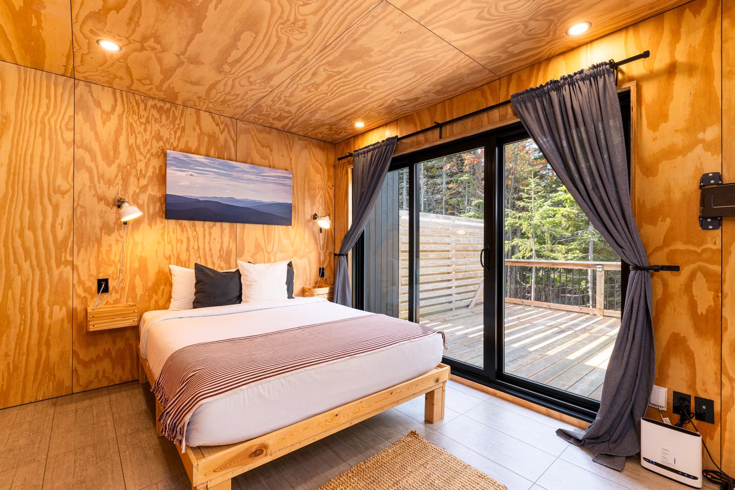 Chambre en bois avec lit double, grandes fenêtres sur terrasse et rideaux gris.
