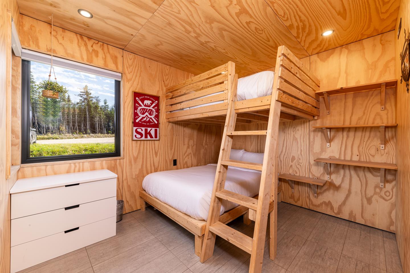 Petite chambre en bois avec lits superposés, commode blanche et fenêtre ouverte sur la forêt.