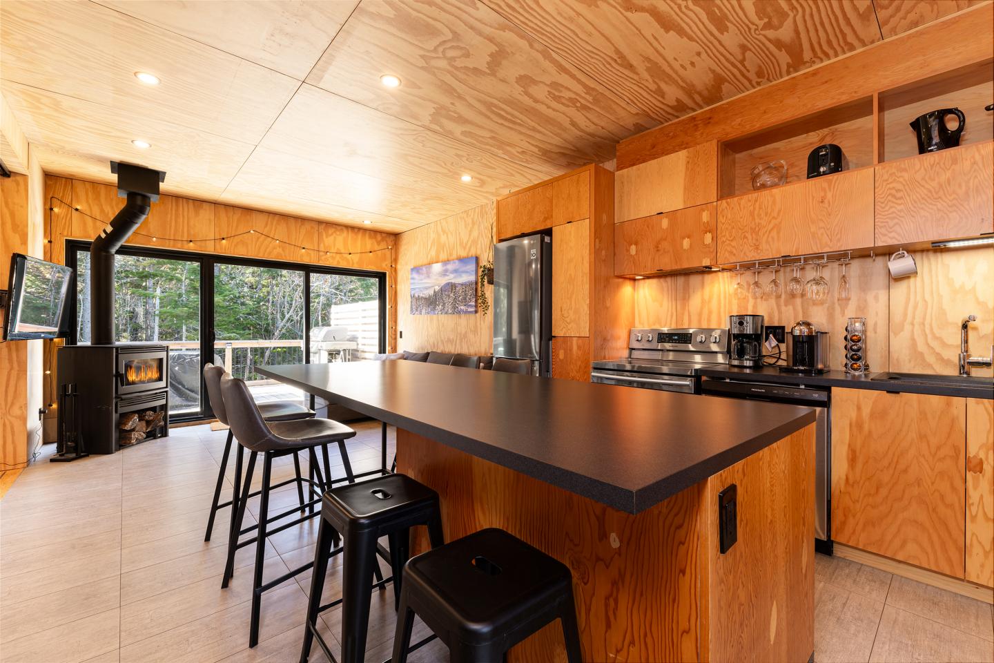 Cuisine moderne en bois avec îlot central, tabourets noirs, et poêle à bois.