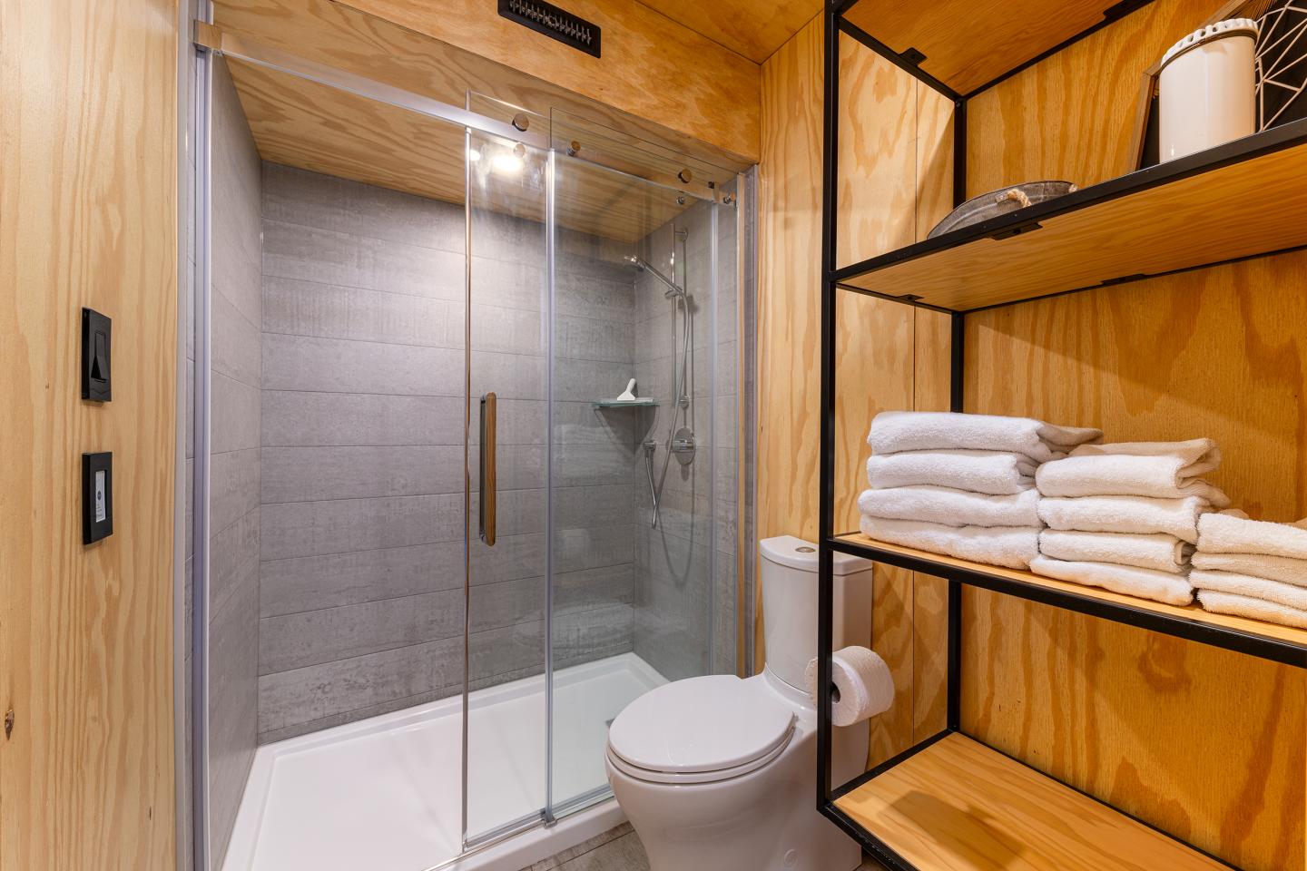 Salle de bain moderne avec douche vitrée, toilettes et étagères en bois avec serviettes.