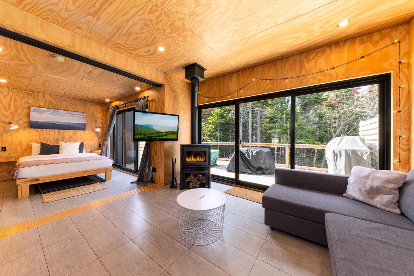Intérieur moderne en bois clair avec lit, canapé, télé, et grande fenêtre donnant sur la forêt.