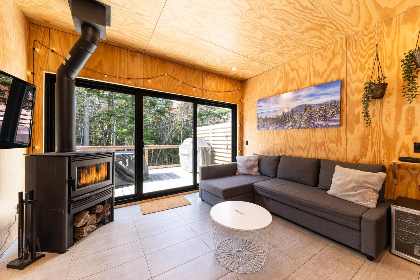 Salon moderne avec poêle à bois, canapé gris et table basse blanche, vue sur terrasse boisée.