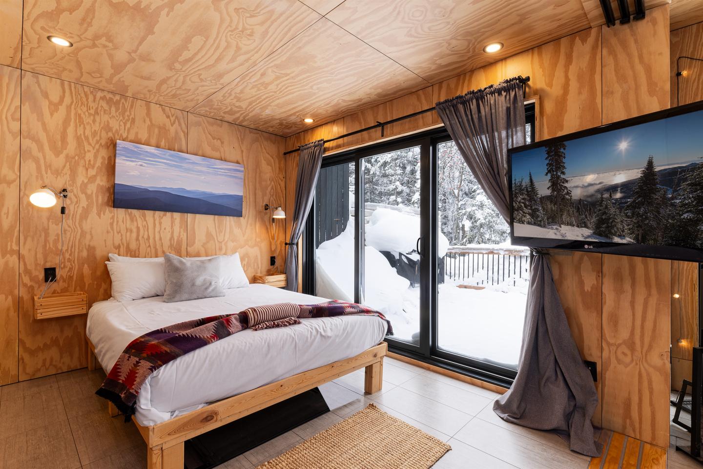Chambre en bois avec lit, tableau, et vue enneigée par la fenêtre.