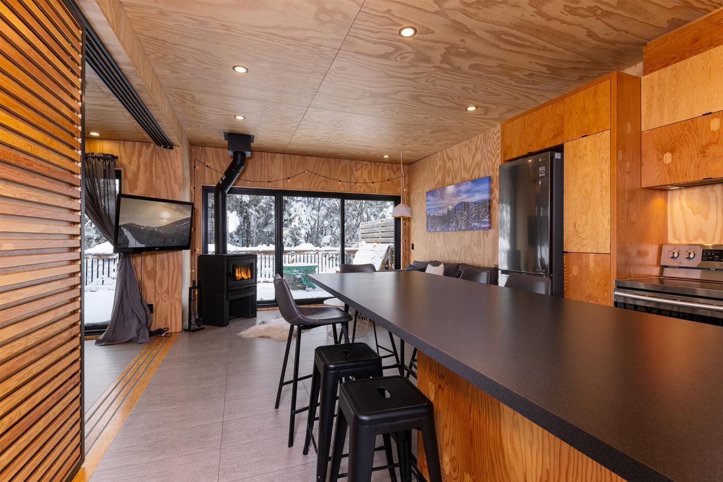Cuisine moderne en bois, avec îlot central, grandes fenêtres et poêle à bois allumé.