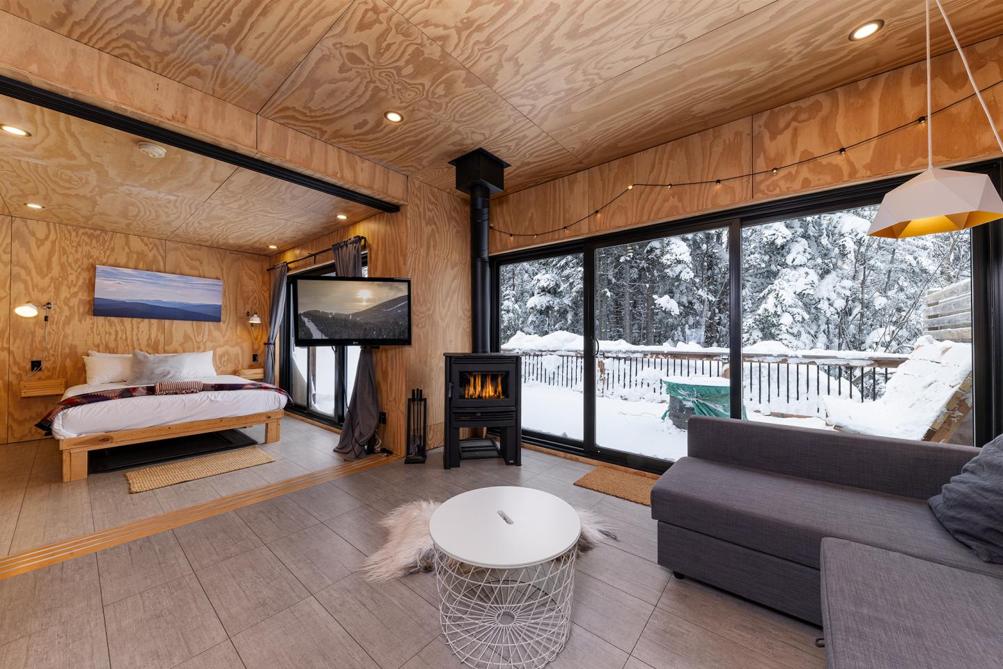 Chalet en bois avec poêle, canapé, et grande fenêtre sur neige.