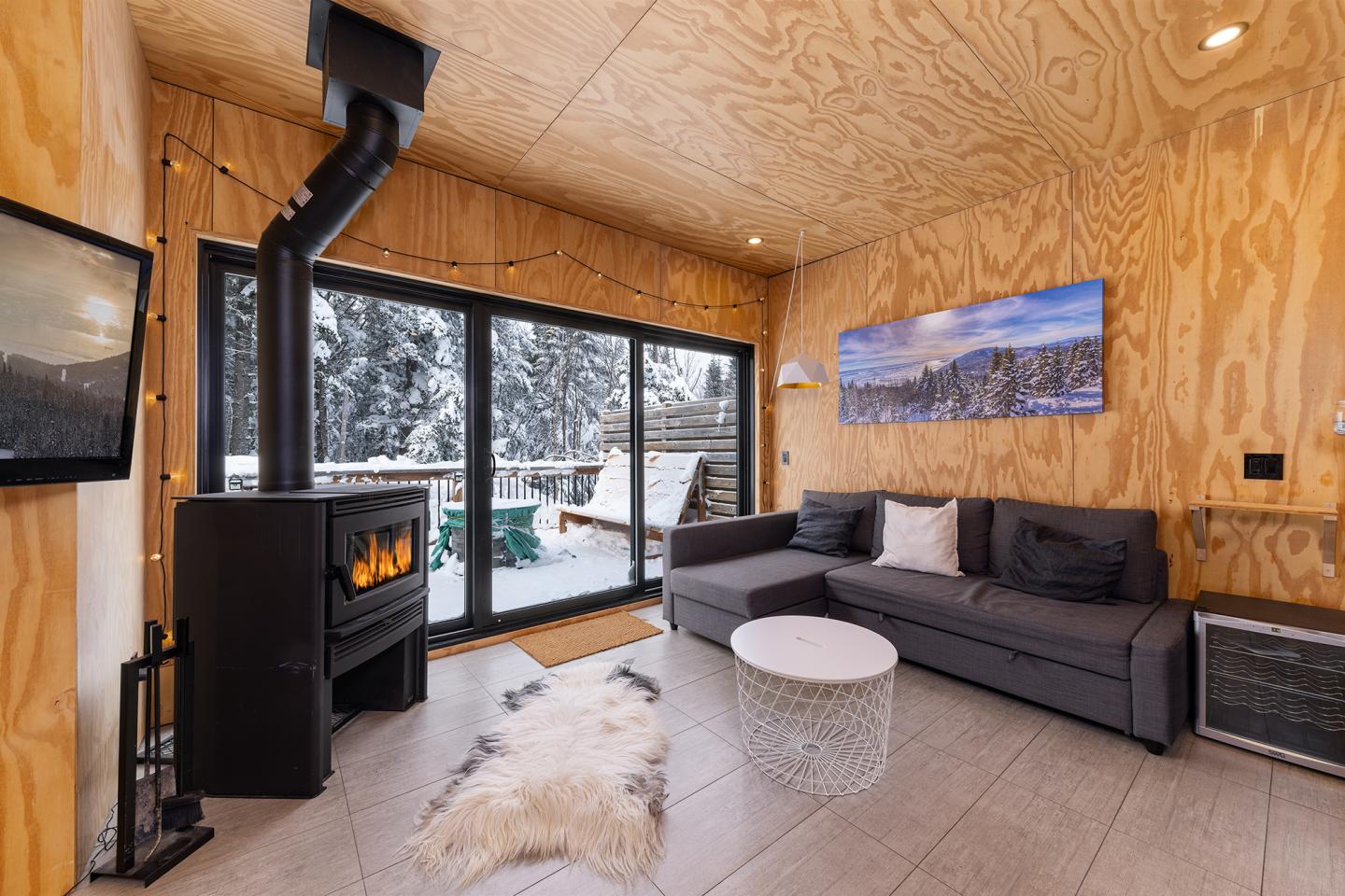 Salon en bois, poêle à bois, canapé gris, vue sur forêt enneigée.