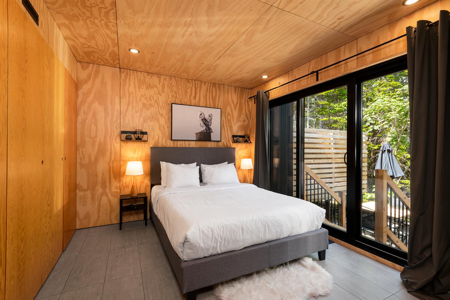 Chambre moderne avec lit double, murs en bois, grandes baies vitrées et rideaux sombres.