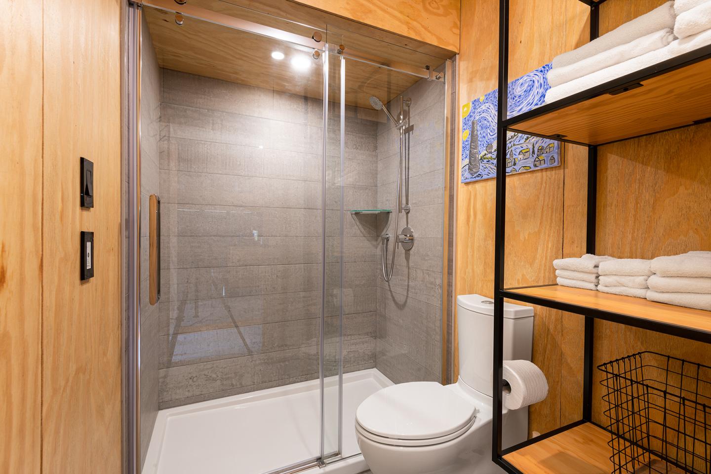Salle de bain moderne avec douche en verre, toilettes, étagères en bois avec serviettes.