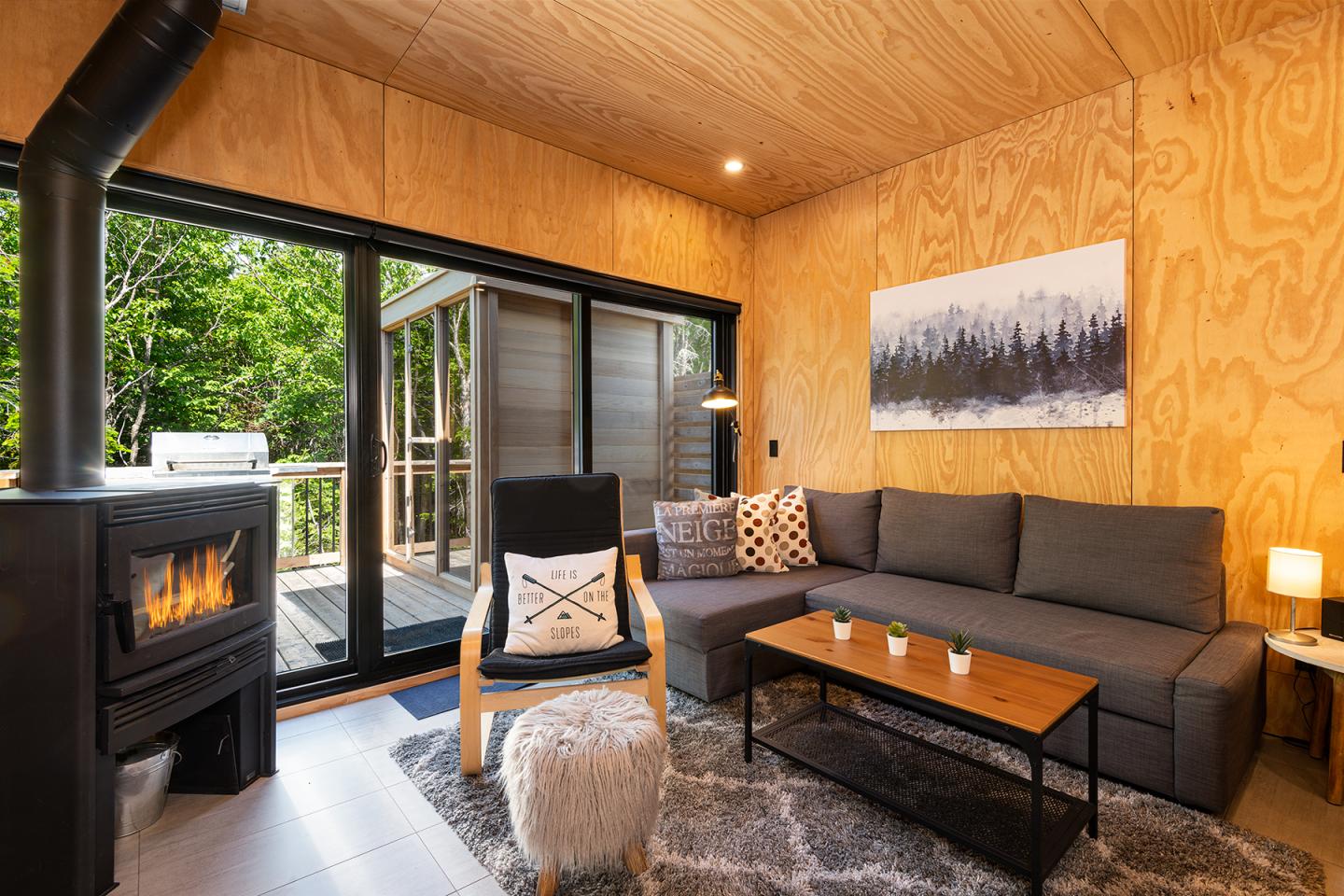 Salon moderne avec canapé gris, poêle à bois, grandes fenêtres et décor boisé.