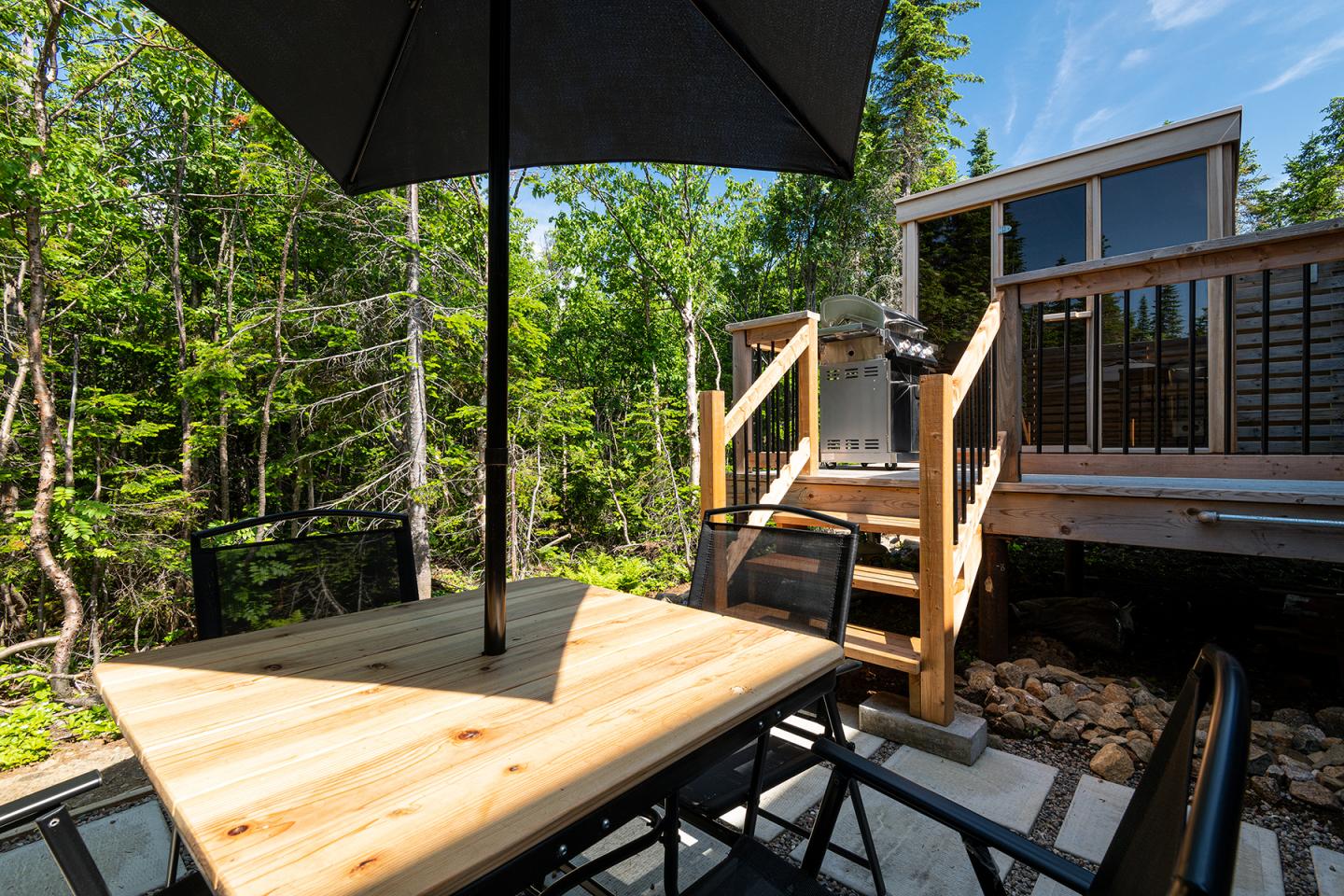 Table en bois sur une terrasse ensoleillée, entourée de forêt verdoyante, avec une maison en arrière-plan.